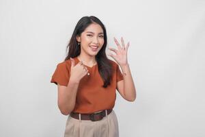 Cheerful Asian woman in brown shirt holding a medicine pill while showing thumbs up gesture isolated by white background. photo