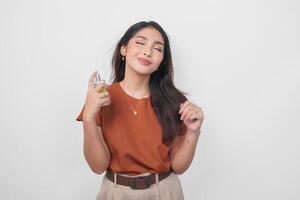 Beautiful young Asian woman in brown shirt holding bottle of perfume and spraying on her neck isolated by white background. photo