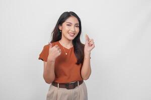 Cheerful Asian woman in brown shirt holding a medicine pill while showing thumbs up gesture isolated by white background. photo