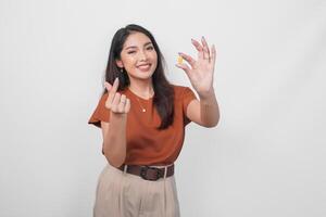 Lovely Asian woman in brown shirt holding a medicine pill while making korean finger heart sign isolated by white background. photo