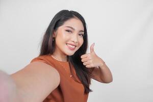 Smiling Asian woman posing for a selfie while giving thumbs up hand gesture of approval, isolated by white background. photo