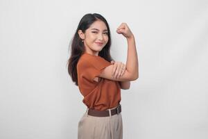 hermosa asiático mujer vistiendo un marrón camisa posando fuerte gesto por levantamiento su brazos y músculos mientras sonriente con orgullo foto