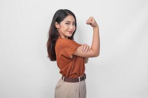 hermosa asiático mujer vistiendo un marrón camisa posando fuerte gesto por levantamiento su brazos y músculos mientras sonriente con orgullo foto