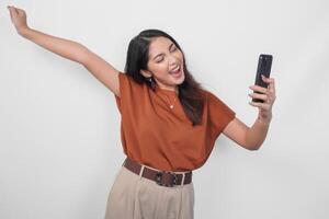 Joyful young Asian woman in brown shirt doing call with happy successful expression and clenched fist gesture isolated over white background. photo