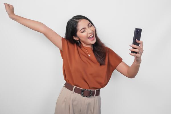 Joyful young Asian woman in brown shirt doing call with happy successful expression and clenched fist gesture isolated over white background.
