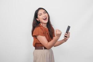 Joyful young Asian woman in brown shirt doing call with happy successful expression and clenched fist gesture isolated over white background. photo