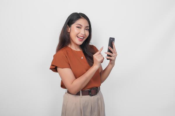 Happy Asian woman is smiling and pointing to her smartphone wearing brown shirt isolated by a white background.