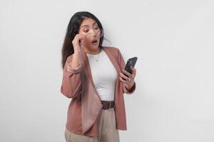 Surprised Asian business woman in eyeglasses looking at shocking news on her phone, isolated by white background. photo