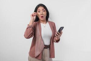 Surprised Asian business woman in eyeglasses looking at shocking news on her phone, isolated by white background. photo