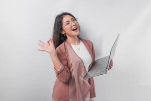 Excited young Asian business woman showing okay sign with right hand and holding laptop on isolated white background. photo