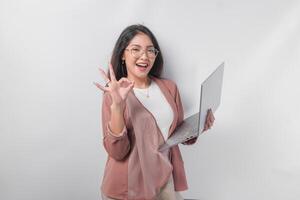 Excited young Asian business woman showing okay sign with right hand and holding laptop on isolated white background. photo