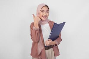 Smiling Asian woman wearing hijab holding document and giving thumbs up hand gesture of approval on isolated white background. photo