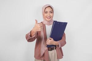 Smiling Asian woman wearing hijab holding document and giving thumbs up hand gesture of approval on isolated white background. photo