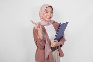 alegre joven musulmán asiático mujer en hijab participación documento mientras señalando a Copiar espacio hacia arriba, aislado por blanco antecedentes. foto