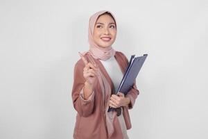 Cheerful young Muslim Asian woman in hijab holding document while pointing to copy space upwards, isolated by white background. photo