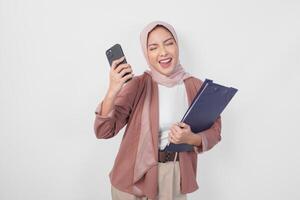 Excited Asian Muslim woman wearing hijab holding a document book and smartphone with cheerful expression over isolated white background. photo