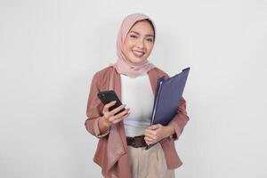 Young Asian Muslim woman wearing hijab holding a document book and smartphone smiling to the camera over isolated white background. photo