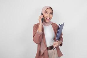 Young Asian Muslim woman wearing hijab talking on a phone call while holding document over isolated white background. photo