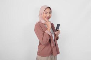 Thoughtful young Asian Muslim woman wearing hijab holding phone while thinking and finding idea with happy expression, isolated on white background. photo