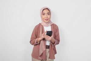 Hungry young Asian Muslim woman wearing hijab looking aside while holding smartphone isolated over white background. photo