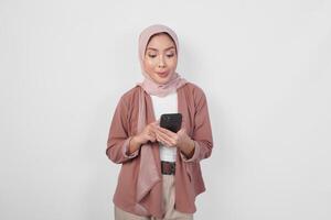 Hungry young Asian Muslim woman wearing hijab looking aside while holding smartphone isolated over white background. photo