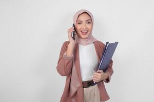 Young Asian Muslim woman wearing hijab talking on a phone call while holding document over isolated white background. photo