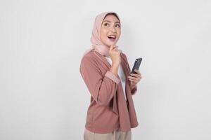 Thoughtful young Asian Muslim woman wearing hijab holding phone while thinking and finding idea with happy expression, isolated on white background. photo