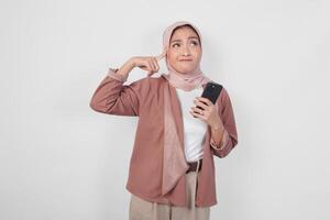 Thoughtful Asian Muslim woman wearing hijab looking aside while holding smartphone isolated over white background. photo