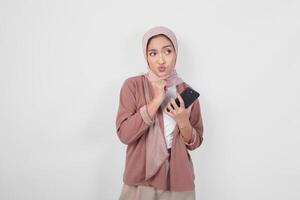 Thoughtful Asian Muslim woman wearing hijab looking aside while holding smartphone isolated over white background. photo
