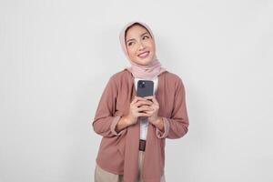 Thoughtful young Asian Muslim woman wearing hijab holding phone while thinking and finding idea with happy expression, isolated on white background. photo