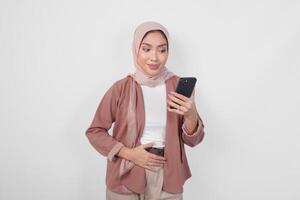 Hungry young Asian Muslim woman wearing hijab looking aside while holding smartphone isolated over white background. photo