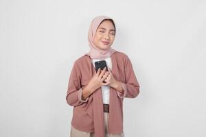 Happy mindful thankful young woman hand on chest smiling while holding smartphone, isolated on white background photo