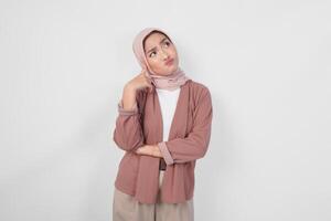 Thoughtful young Asian Muslim woman wearing hijab, holding her chin and thinking with serious expression over isolated white background. photo