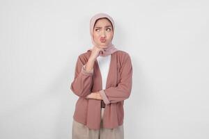 Thoughtful young Asian Muslim woman wearing hijab, holding her chin and thinking with serious expression over isolated white background. photo