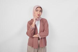 Thoughtful young Asian Muslim woman wearing hijab, holding her chin and thinking with serious expression over isolated white background. photo