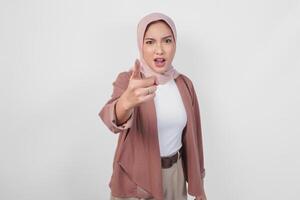 Young Asian Muslim woman wearing hijab looking disgruntled irritated expression while pointing to camera, isolated by white background. photo