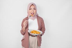 Hungry young Asian Muslim woman in hijab holding fork and plate eating french fries over isolated white background. photo