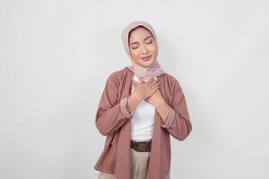 Happy mindful thankful young Asian Muslim woman wearing hijab hands on chest smiling isolated on white background. photo