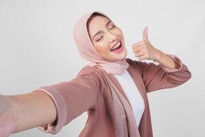 Smiling Asian woman in hijab posing for a selfie while giving thumbs up hand gesture of approval, isolated by white background. photo