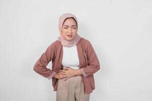 An unwell sick young Asian Muslim woman placing hand on stomach suffers pain isolated on white color background. photo