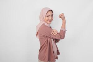 Excited Asian Muslim woman wearing a hijab showing strong gesture by lifting her arms and muscles smiling proudly. photo