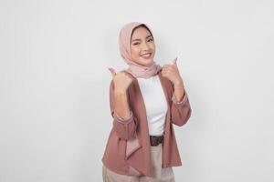 Smiling Asian Muslim woman wearing a hijab giving thumbs up hand gesture of approval, isolated by white background. photo