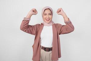 Cheerful young Asian Muslim woman in hijab doing a successful win gesture with clenched fist over isolated white background. photo
