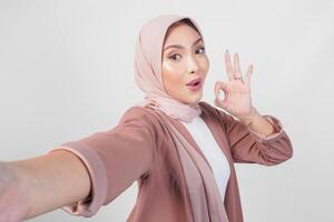 Happy Asian Muslim woman in hijab doing selfie pose giving an okay hand gesture isolated over white background. photo