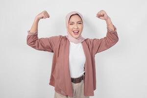 Excited Asian Muslim woman wearing a hijab showing strong gesture by lifting her arms and muscles smiling proudly. photo