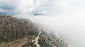 Top view of coastal mountains with city and clouds. Action. Dense white clouds approach mountains near coast. Coastline with mountains and city is covered with dense clouds from sea video