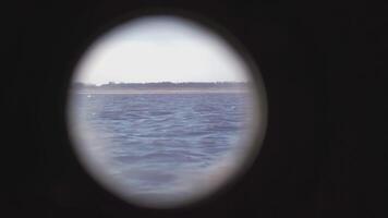 View from the round porthole of the ship to the shore and waves. CLIP. Landscape on the shore covered with trees and the sea with waves from the porthole of the ship video