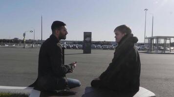 jung männlich freunde Sitzung auf ein Bank beim Bus halt und reden. hdr. zwei Männer warten zum ein Bus draußen während Konversation auf ein sonnig Tag. video