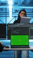 Vertical Multiethnic engineers working in high tech server hub, using green screen laptop and PCs to analyze data. Diverse employees examining infrastructure in data center, using chroma key notebook video