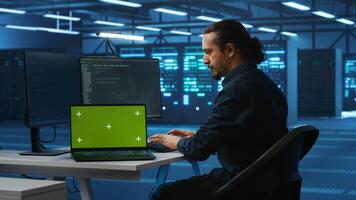 Engineer upgrading equipment in data center using green screen laptop. Specialist at PC desk in high tech establishment overseeing server rows with mockup notebook, panning shot video